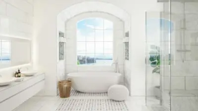 Hvidt badeværelse med hvidt badekar og hvidt flisegulv og hvide vægge foran et stort lyst vindue der har en udsigt til havet