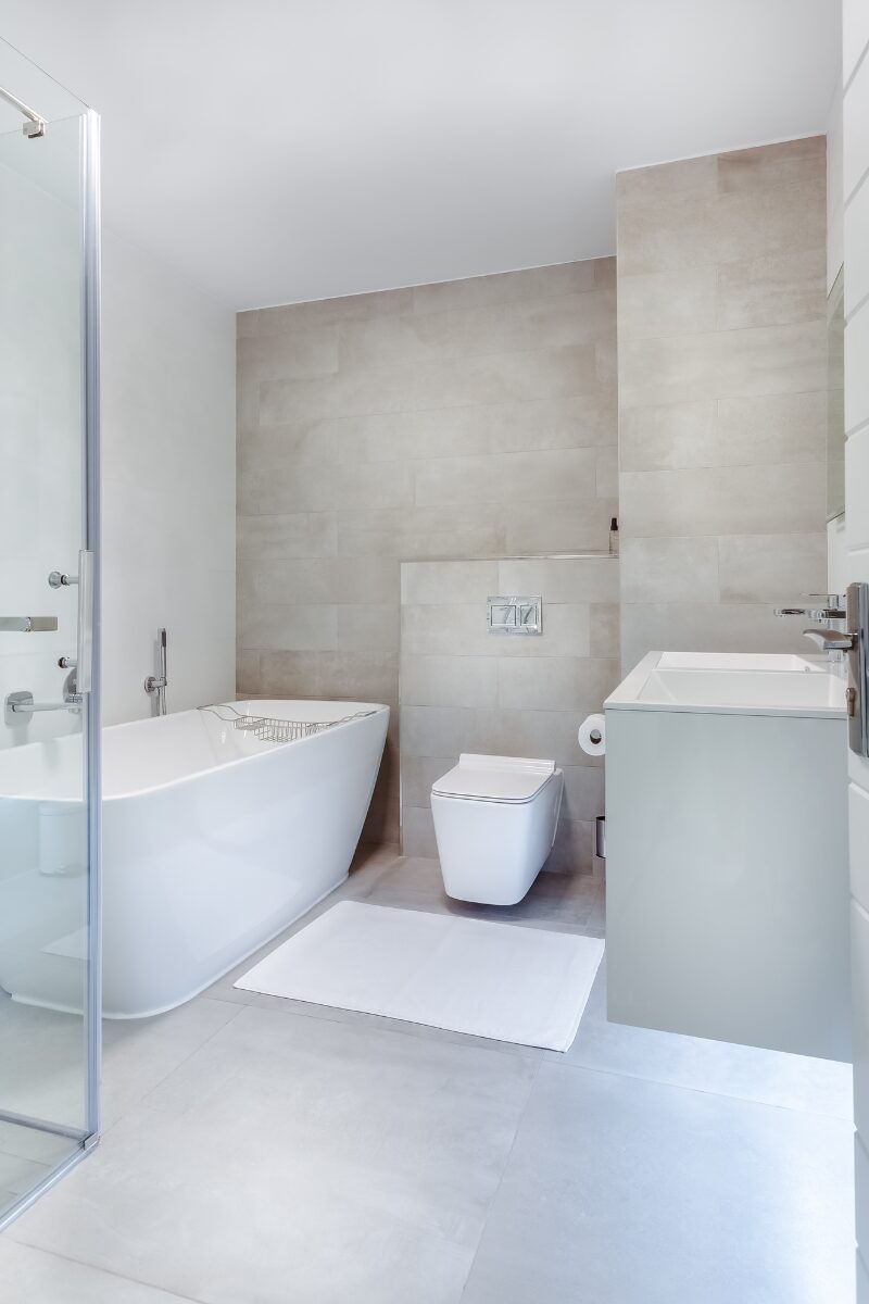 moderne og lyst badeværelse med med hvide og beigefarvet vægge. Der er en brusekabine af glas, en håndvask, et hvidt badekar og et toilet. 