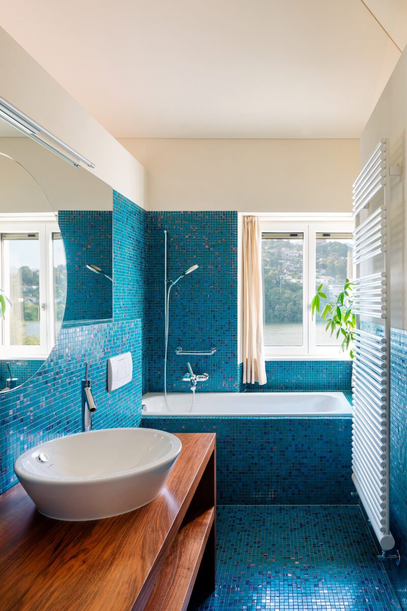 Badeværelse med turkisblå fliser på vægge og gulv. Der en hvid håndvask på et træbord i forgrunden og et hvidt vindue der leder ud til en have i baggrunden. 