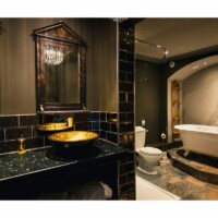 Luksuriøst badeværelse med sort badeværelsesbord i forgrunden med 2 håndvaske i guld. Håndvaskene står foran 2 spejl med rammer i mørkt træ. I baggrunden er der et hvidt badekar med ben bagved et hvidt toilet.