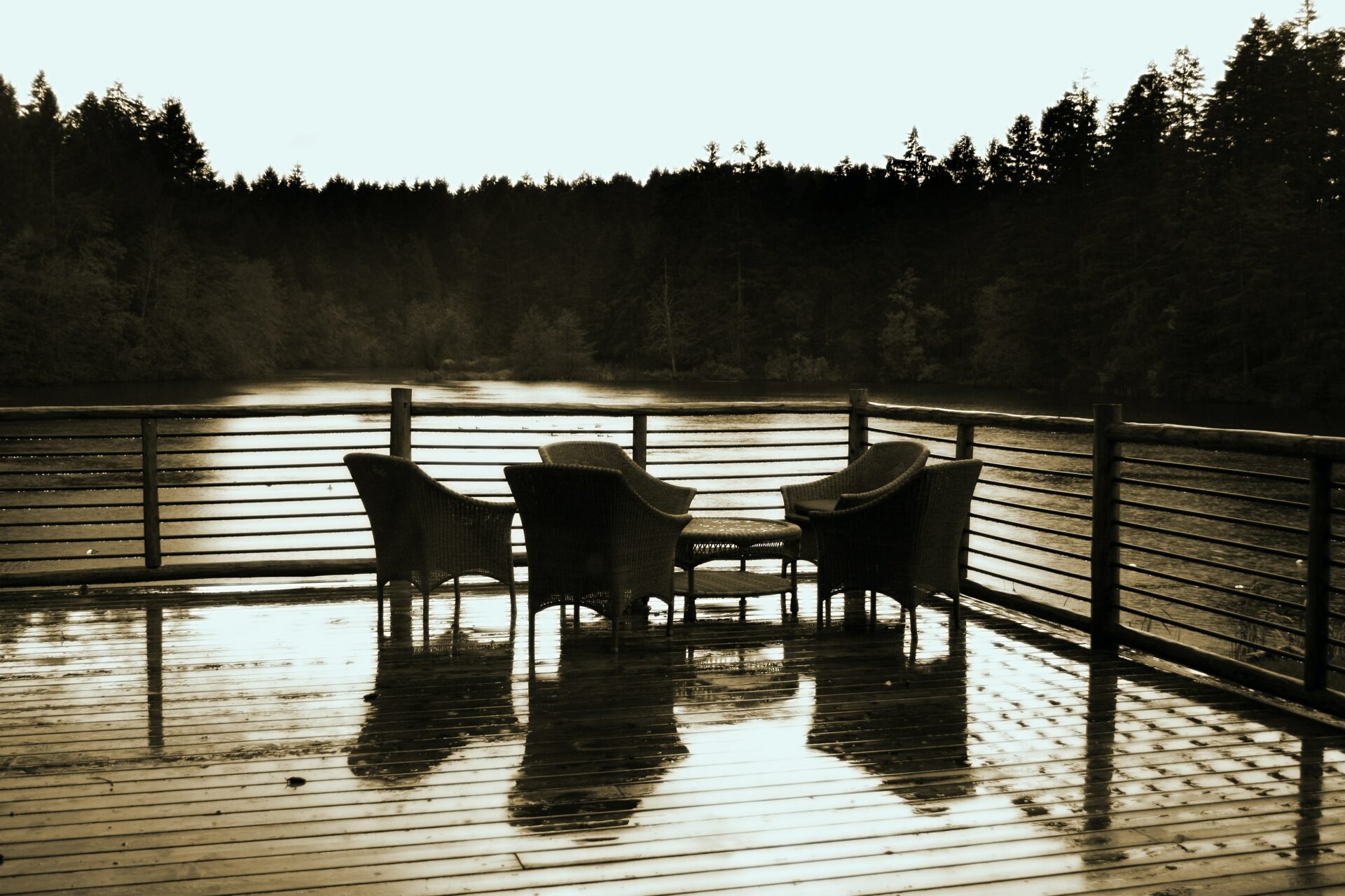 træterrasse ud til vandet med træborde og træstole der står foran et træhegn under en solnedgang der skinner ned på naturen