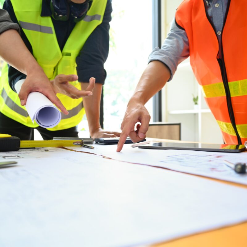 Tre byggeleder står om et bord med arkitekttegninger og diskuterer 